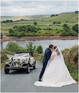 couple kiss at Digley Reservoir with wedding car in the background and green landscape behind them.