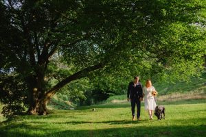 man and woman in wedding attire walk towards the camera by a huge tree showing the landscape behind them