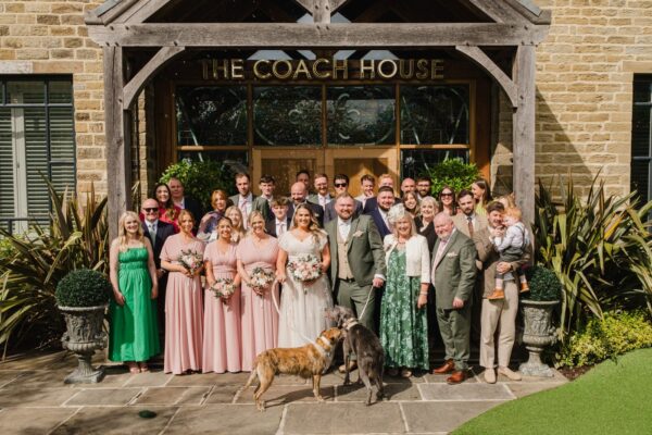 lareg group of wedding guests outside the Coach House at Manor House wedding venue. People smiling and two dogs at the front of the group