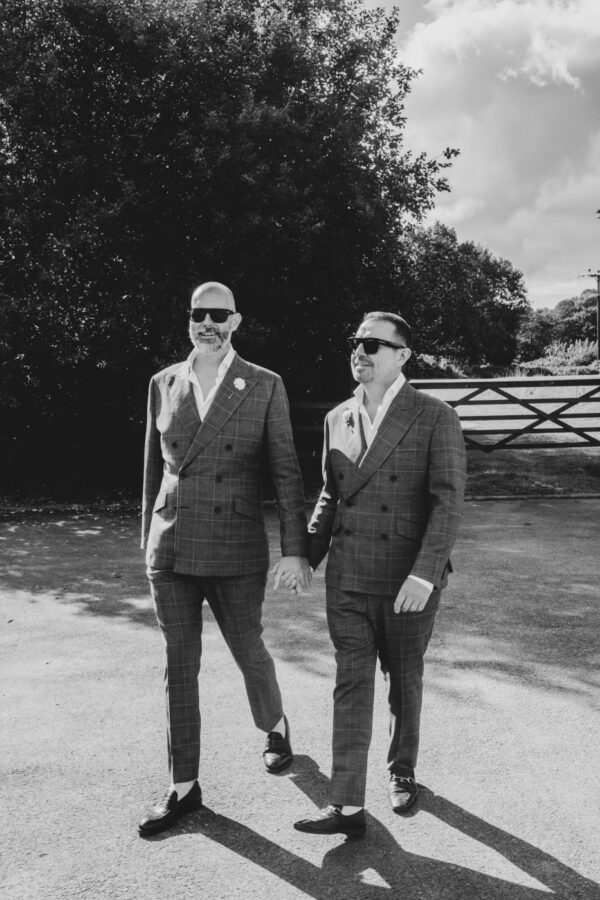 Two grooms walk with sun behind them at the wedding venue (The Old Barn) after their ceremony. Black and White photo, matching suits and sunglasses. Sunny day with long shadows